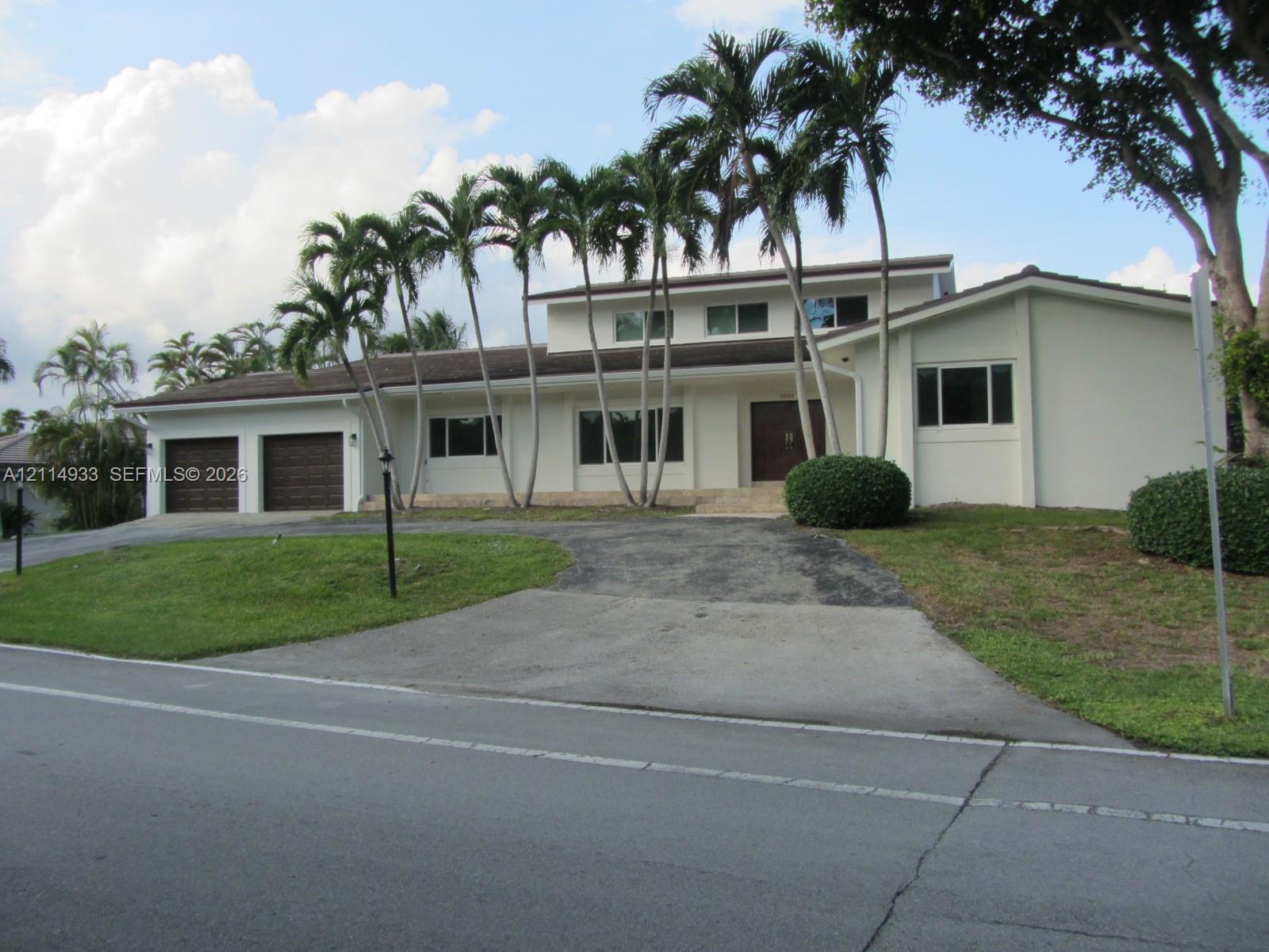 Two-story residence featuring a stucco exterior, multi-car garage with wood-finish doors, expansive driveway, and mature palm tree landscaping
