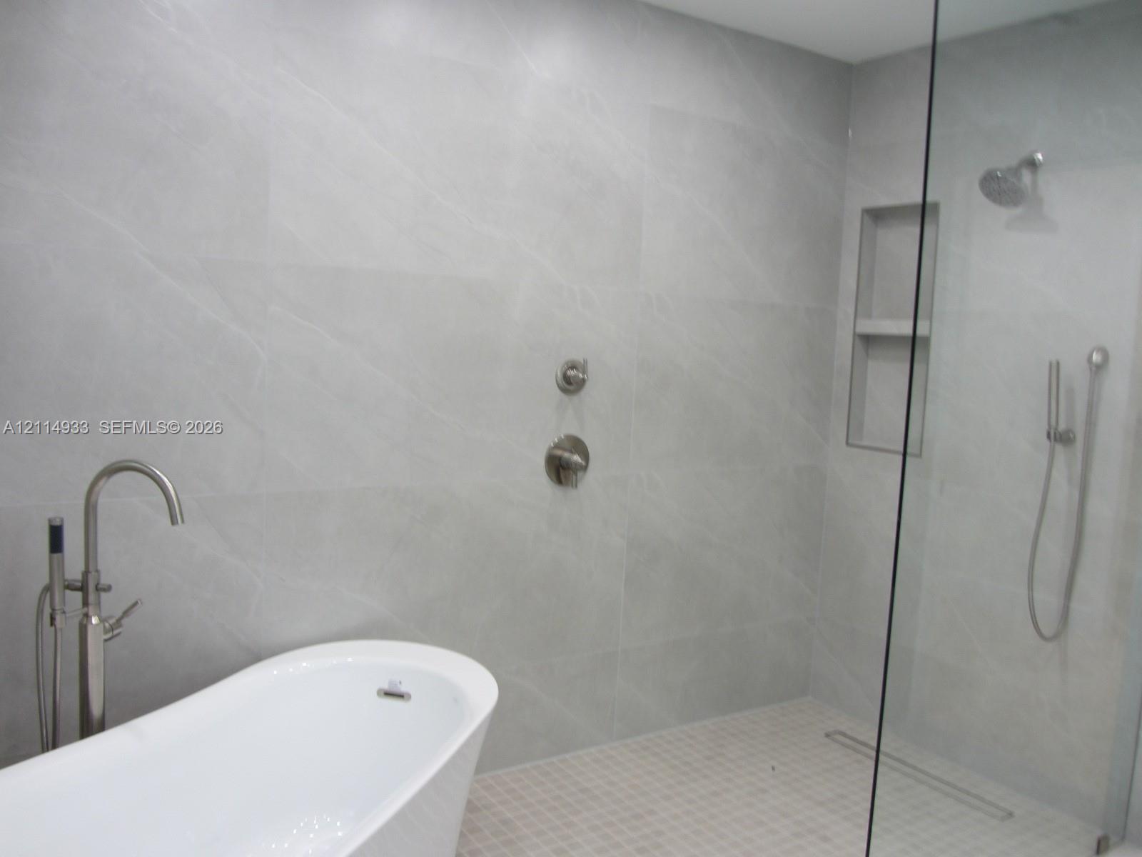 Freestanding soaking tub with floor-mounted faucet, walk-in shower featuring large-format tile surround, integrated tiled wall niche, rain shower head with handheld attachment, and seamless glass partition