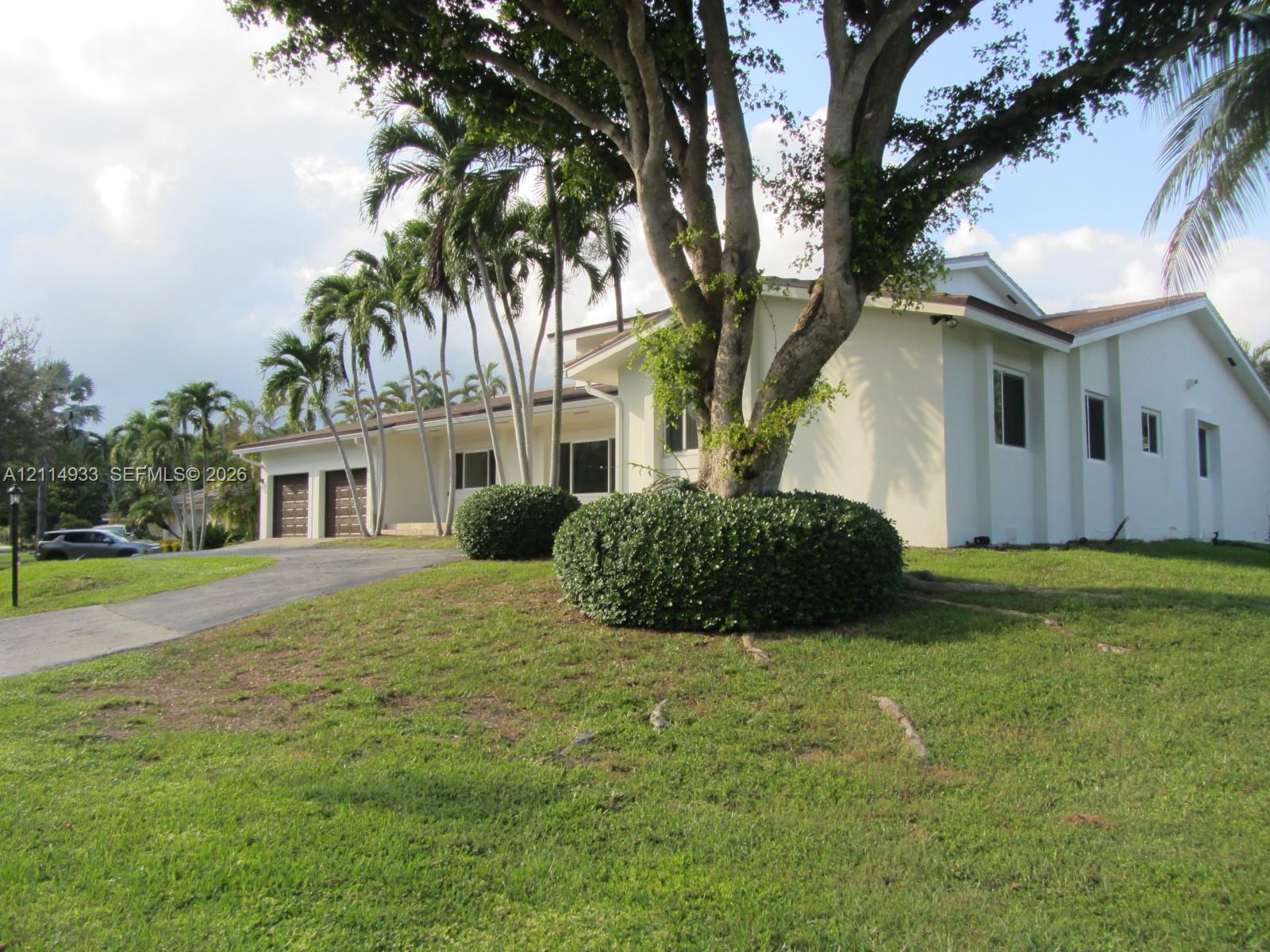 Stucco exterior featuring a two-car garage, expansive lawn, mature shade trees, and ornamental palms Stucco exterior featuring a two-car garage, expansive lawn, mature shade trees, and ornamental palms