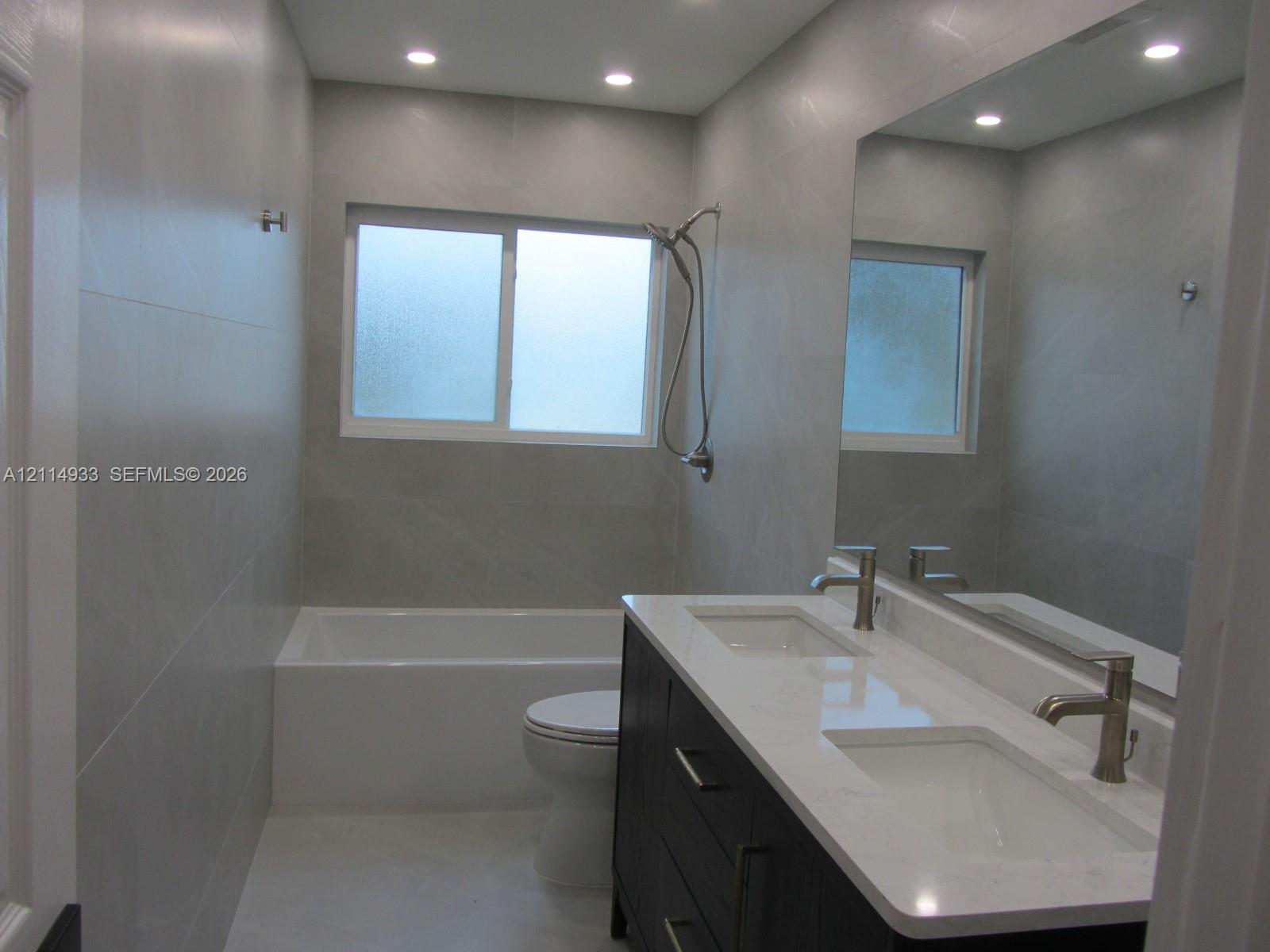 Dual-sink vanity with a quartz countertop, full-width wall mirror, recessed ceiling lighting, and large-format tile wall surround with a built-in tub