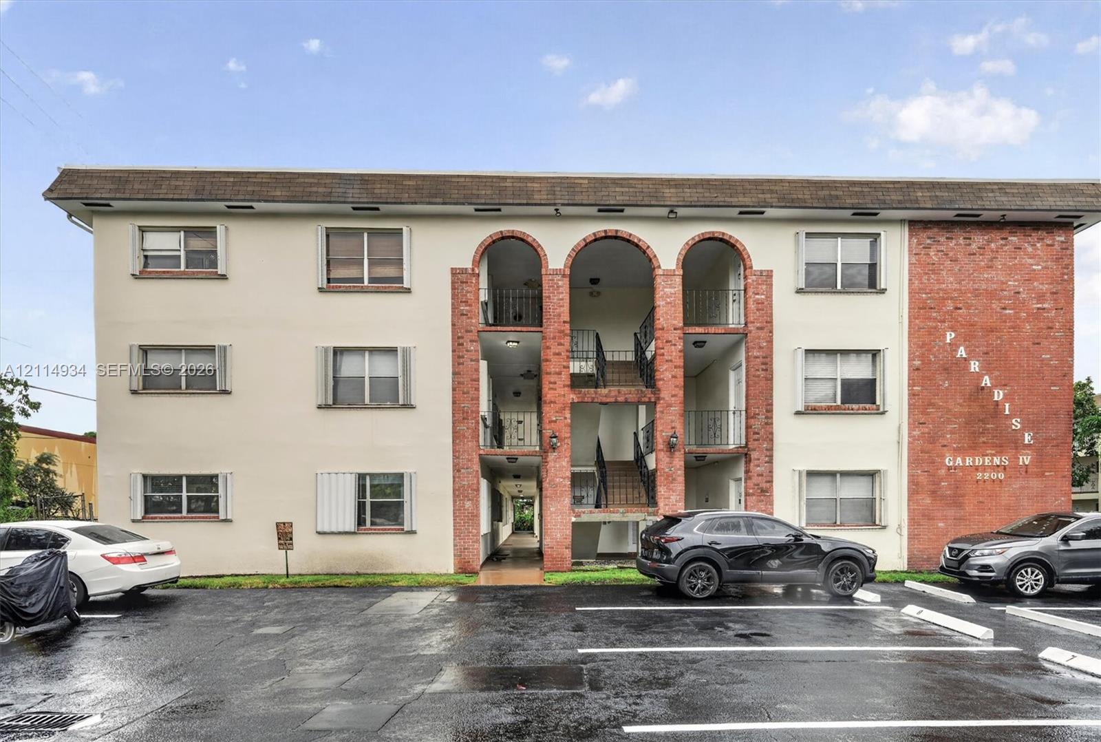 Exterior facade featuring a combination of stucco and red brick masonry, arched entryway detailing, metal balcony railings, and surface parking