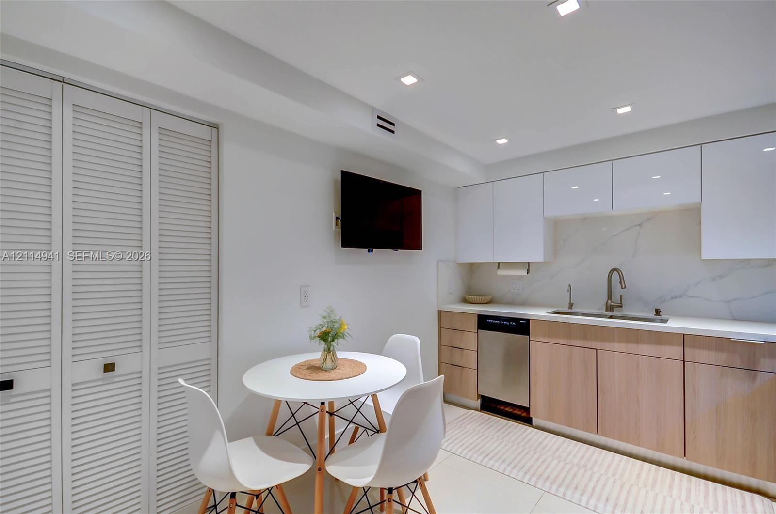 Kitchen area featuring high-gloss white upper cabinetry, wood-finish lower cabinetry, stone-style slab backsplash, recessed ceiling lighting, and floor-to-ceiling louvered closet doors