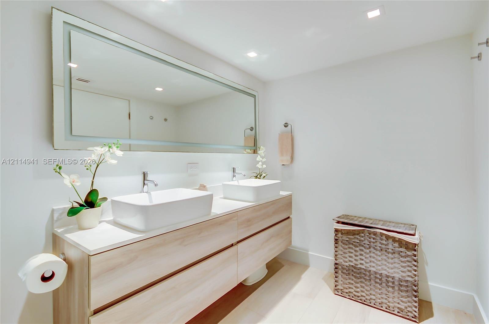 Floating dual-vanity cabinet with light wood-finish cabinetry, white countertop, rectangular vessel sinks, and an oversized illuminated wall mirror with integrated lighting
