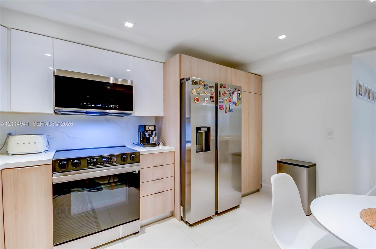 Kitchen featuring high-gloss white upper cabinetry, wood-finish base cabinetry, stone-surface backsplash, stainless steel appliances, and recessed ceiling lighting