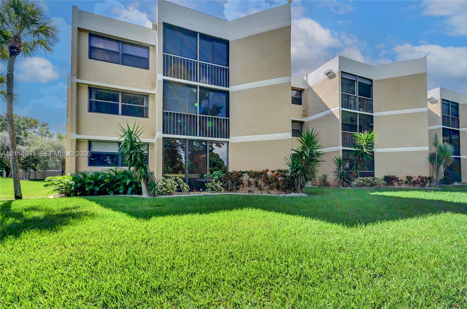 Multi-story exterior featuring a neutral stucco facade, stacked balconies with metal railings, expansive glass windows, and mature perimeter landscaping