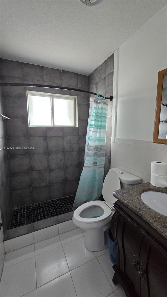 Bathroom featuring a walk-in shower with charcoal-toned tile surround and dark tiled flooring, a vanity with a stone-finish countertop, and white tile flooring
