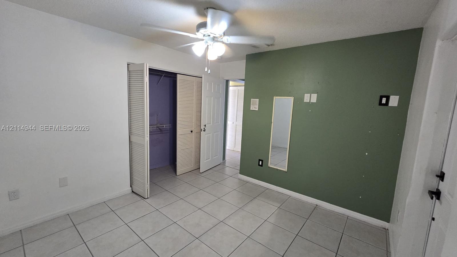 Neutral-toned ceramic tile flooring, ceiling fan with integrated lighting, closet with louvered bi-fold doors, and a decorative accent wall
