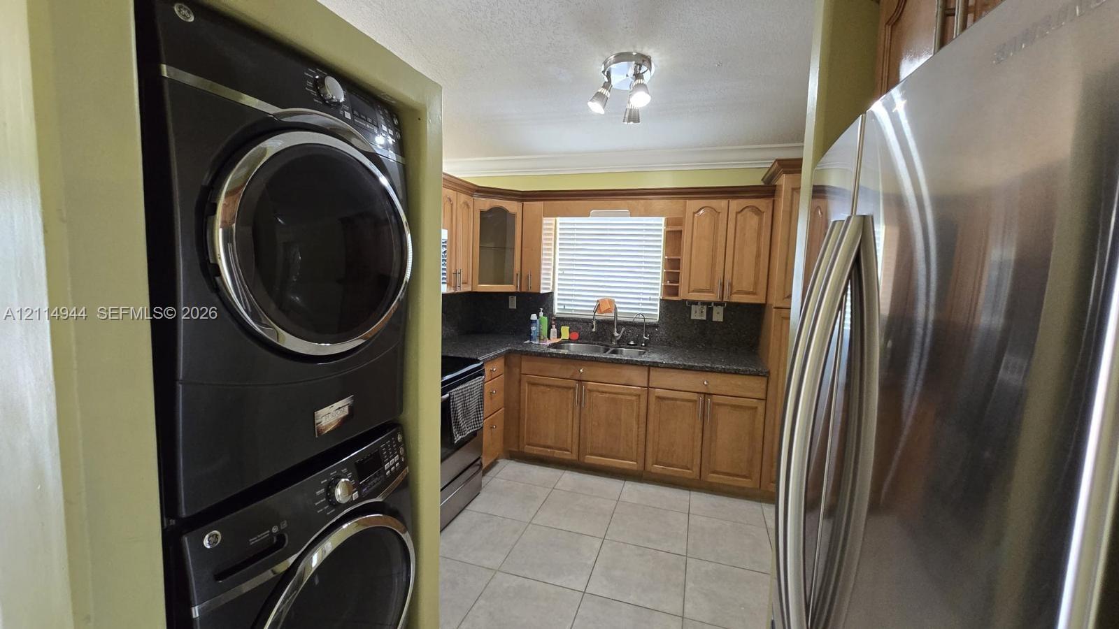 Kitchen featuring medium-tone wood cabinetry, dark stone-finish countertops, stainless steel side-by-side refrigerator, stackable laundry unit, and ceramic tile flooring