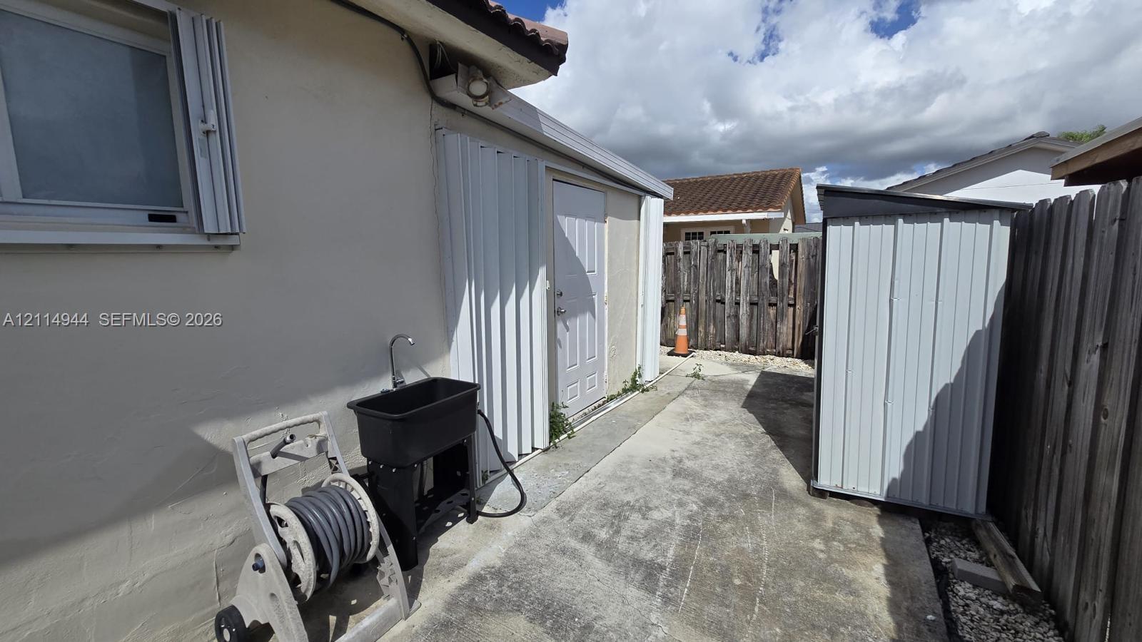 Exterior side yard featuring a concrete surface, a built-in utility sink, accordion-style storm shutters, and an attached metal storage unit enclosed by wood privacy fencing