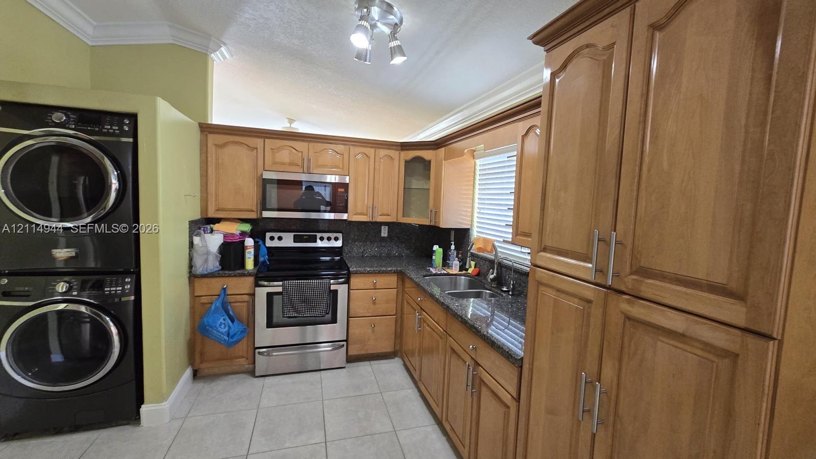 Kitchen featuring natural wood cabinetry, stone-finish countertops, stainless steel appliances, tile flooring, and integrated laundry area