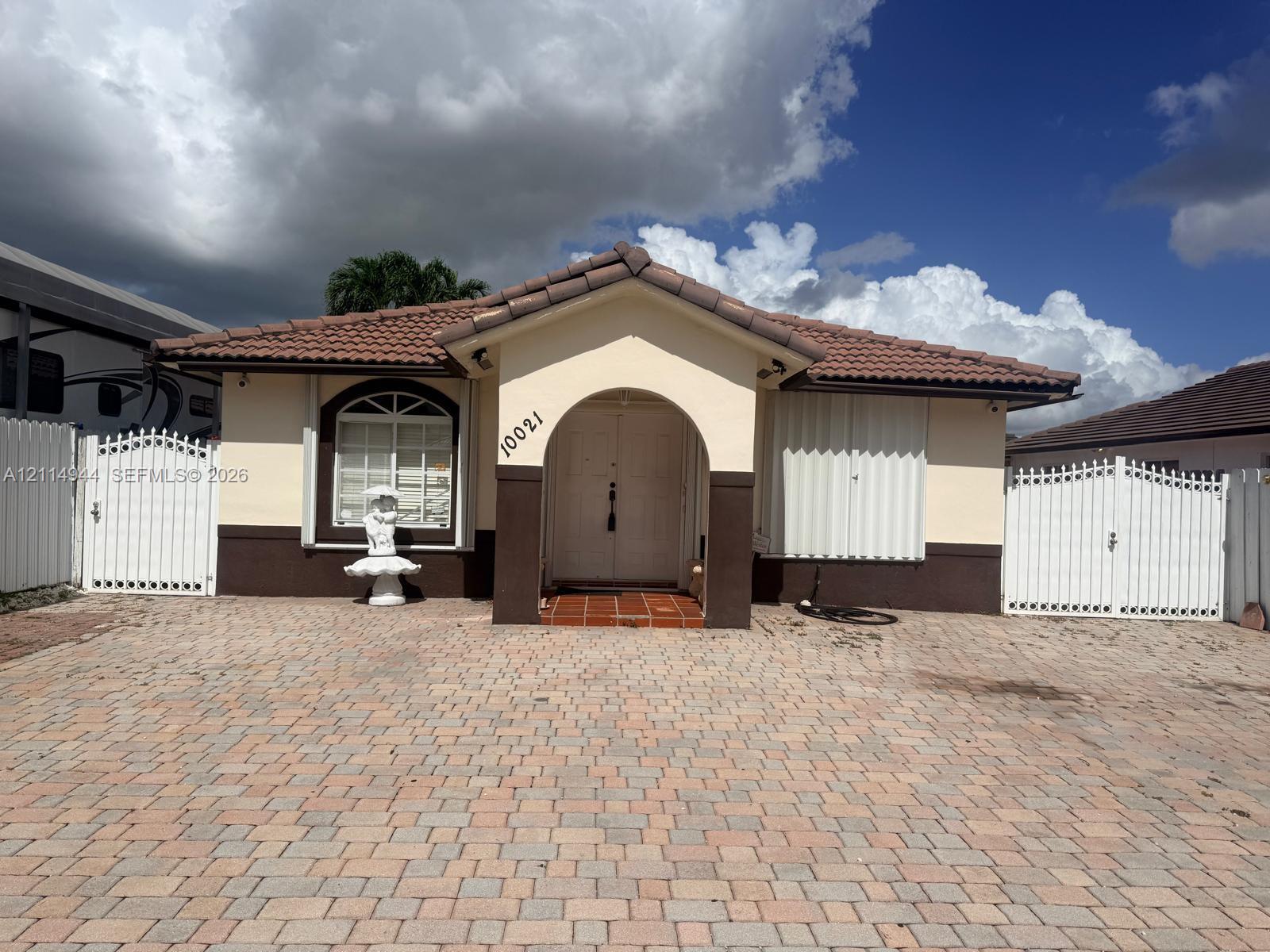 Stucco exterior featuring a tiled roof, arched entryway, brick-paver driveway, and dual gated perimeter fencing