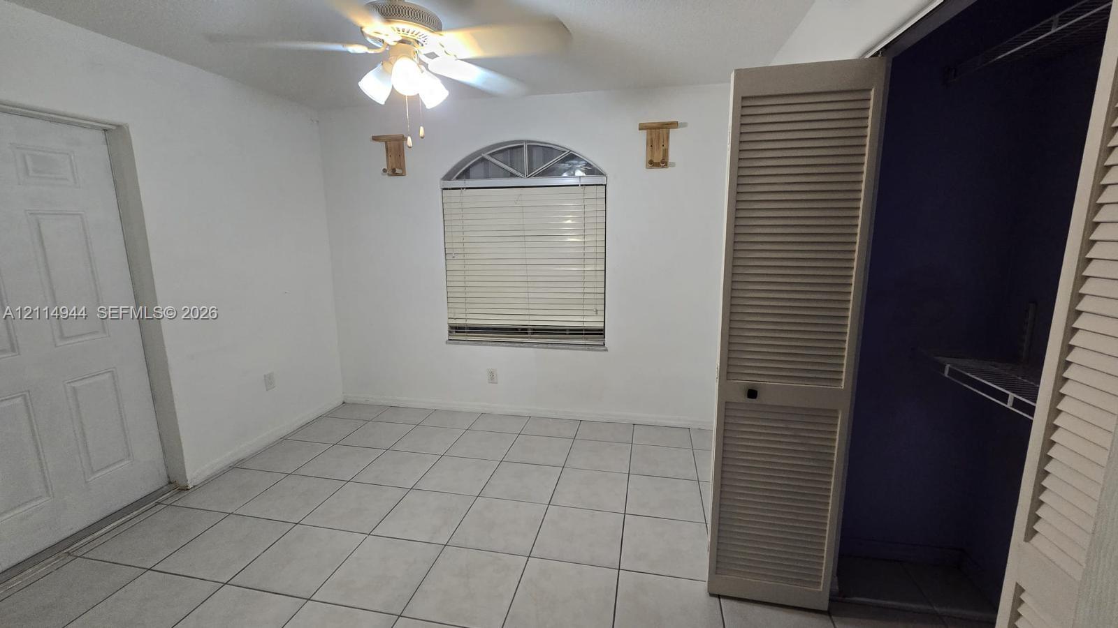 Interior space featuring ceramic tile flooring, arched window with horizontal blinds, ceiling fan with integrated lighting, and louvered closet doors