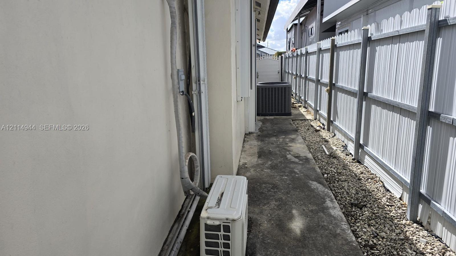 Side exterior featuring a concrete walkway, light-colored stucco siding, a metal privacy fence, and an exterior air conditioning unit