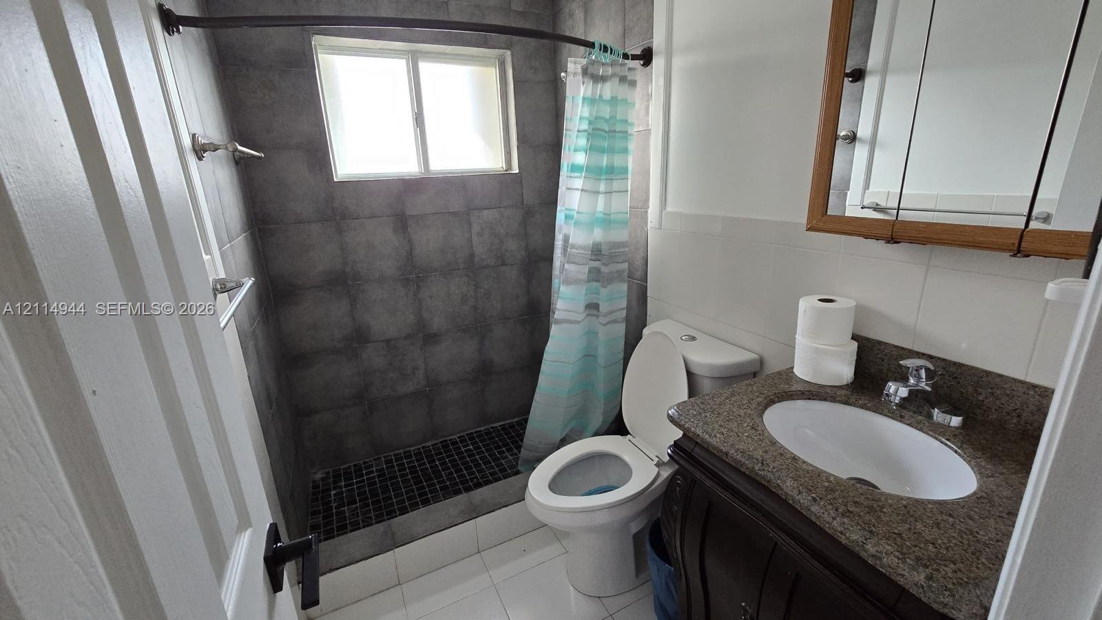 Bathroom featuring a walk-in shower with charcoal-toned wall tiles, dark mosaic floor tiling, stone-surfaced vanity with an integrated basin, and a wood-finish medicine cabinet