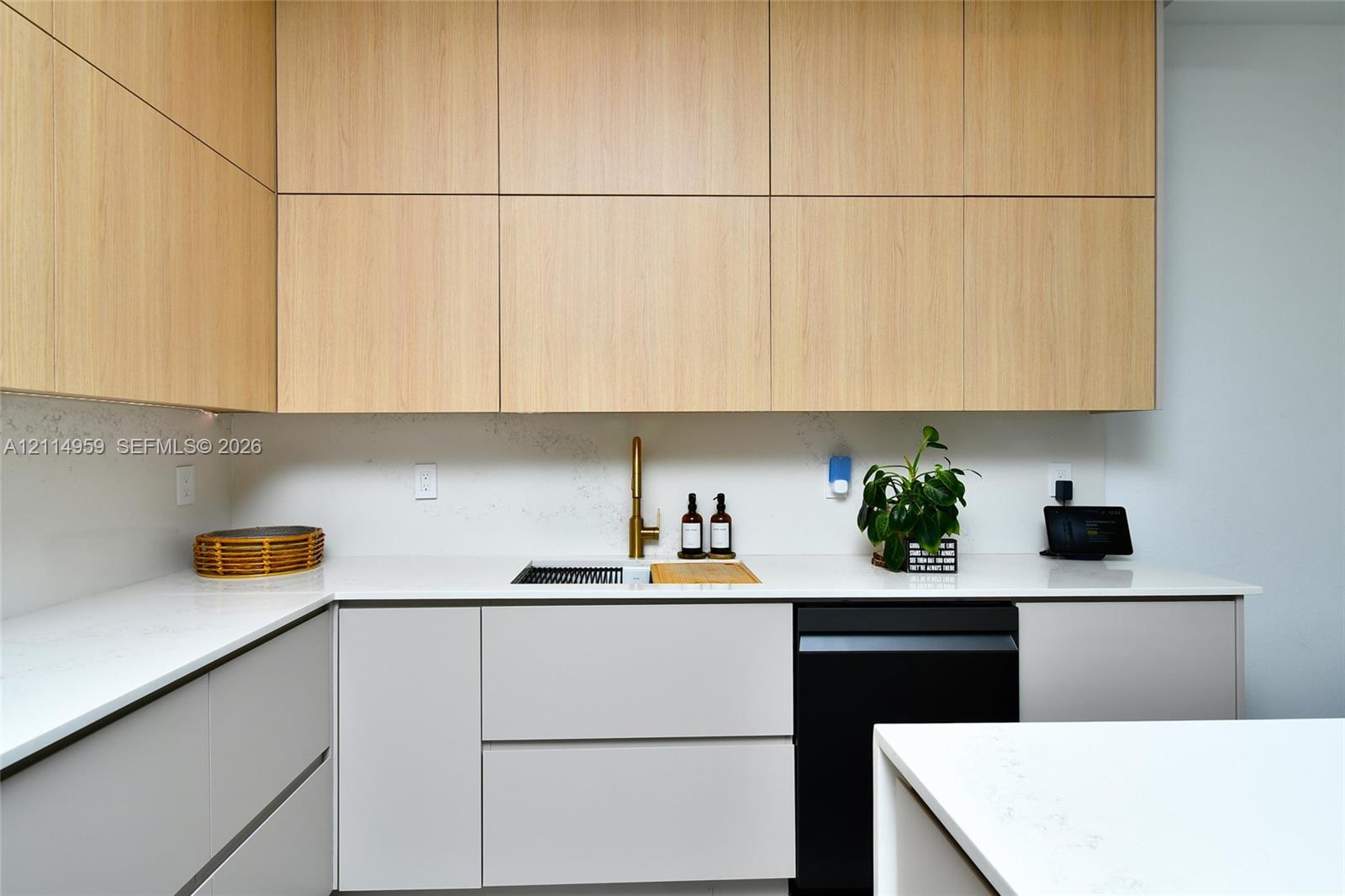 Light-toned wood-finish cabinetry, sleek matte-finish lower cabinetry, stone-composite countertops, integrated workstation sink with metallic faucet, and matte-black dishwasher