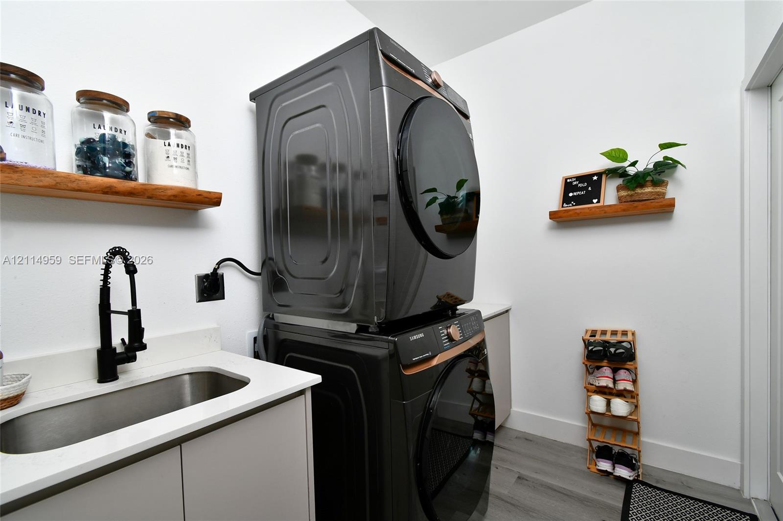 Stacked laundry appliances featuring a metallic finish, undermount stainless steel sink, matte black gooseneck faucet, quartz-surfaced countertop, and natural wood-finish wall-mounted shelving