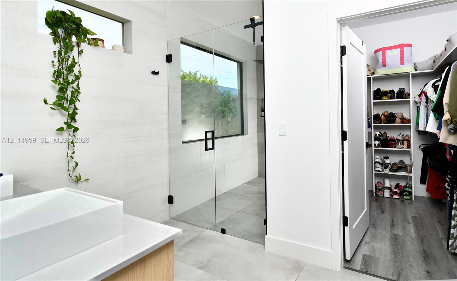 Bathroom featuring a frameless glass-enclosed shower with matte black hardware, large-format tile flooring, rectangular vessel sink on a stone-topped vanity, and a walk-in closet with built-in shelving and wood-finish flooring