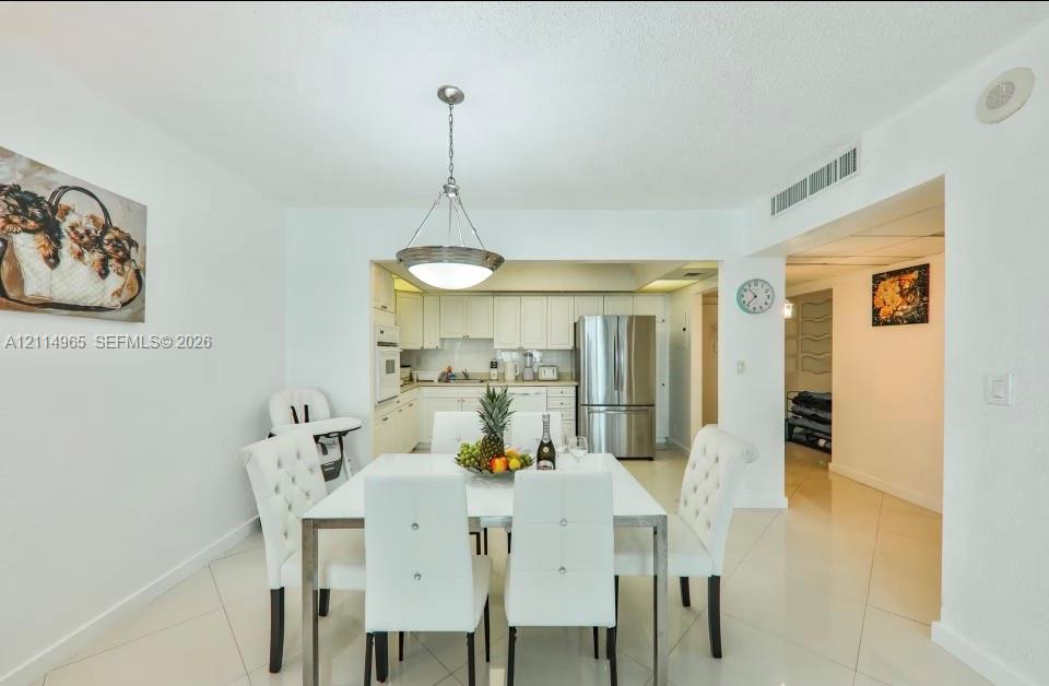 Dining area featuring large-format ceramic tile flooring and a polished metal pendant light fixture, extending to a kitchen with white cabinetry and stainless steel refrigerator