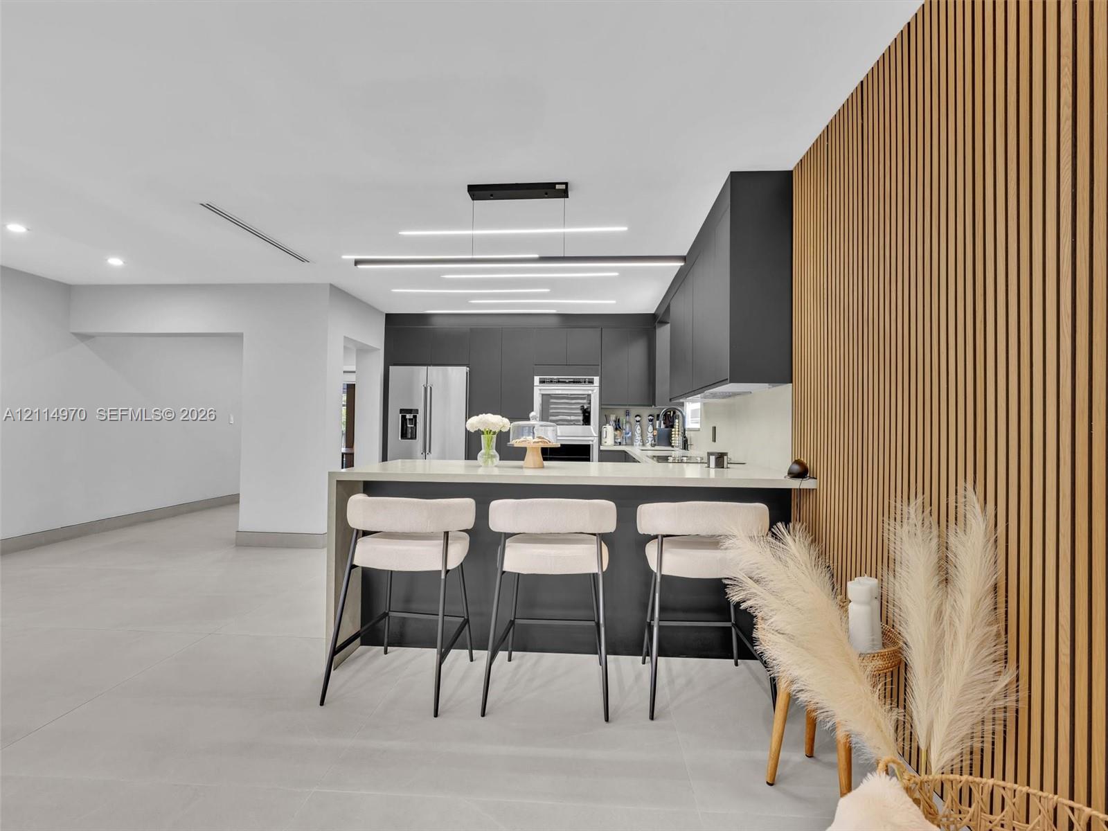 Kitchen featuring matte black flat-panel cabinetry, a waterfall-edge island with stone-surface countertop, floor-to-ceiling slatted wood wall accent, and contemporary linear LED lighting