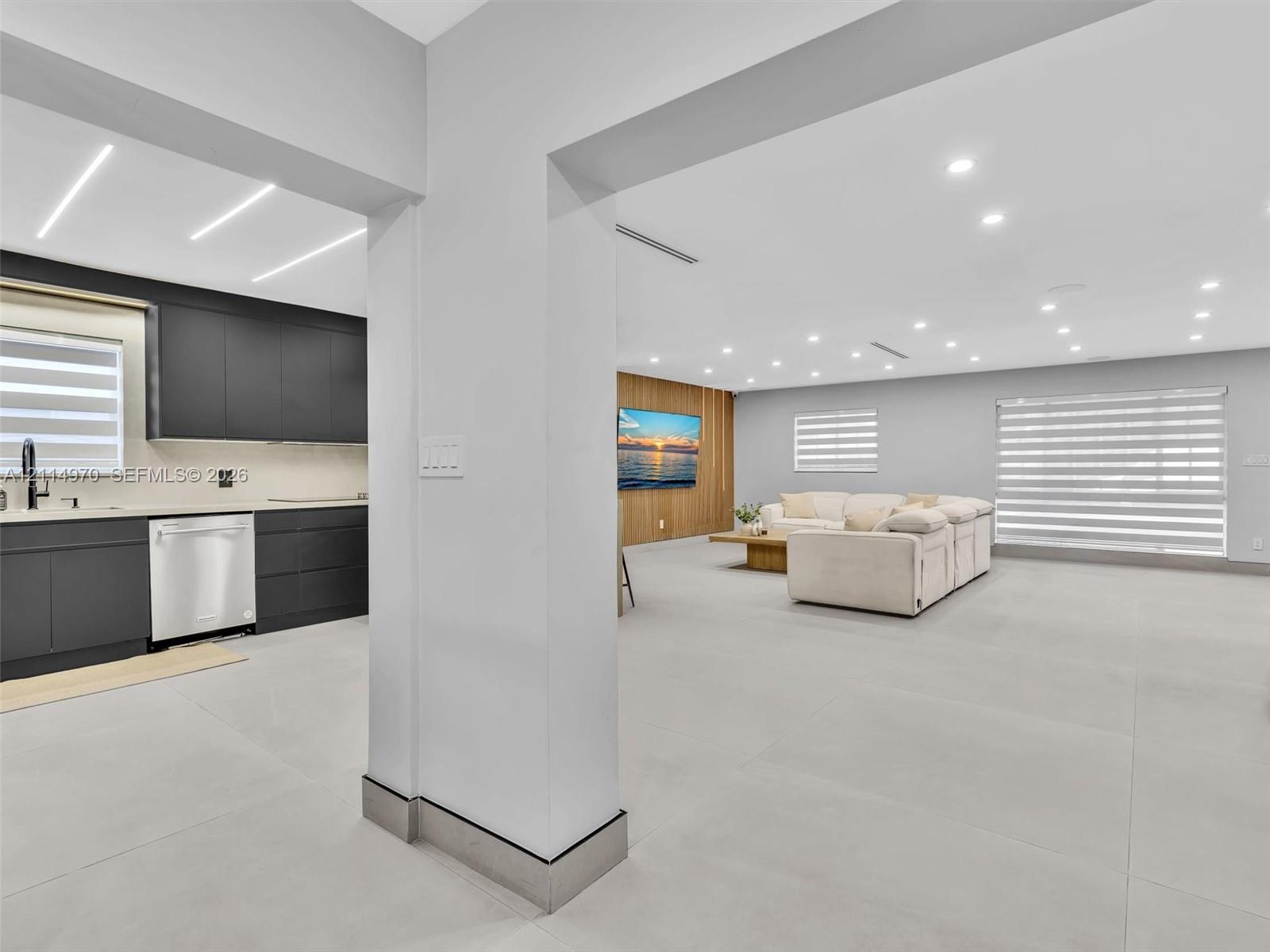 Open-concept interior featuring large-format tile flooring, matte charcoal cabinetry, recessed ceiling lighting, and a wood-slat accent wall Open-concept interior featuring large-format tile flooring, matte charcoal cabinetry, recessed ceiling lighting, and a wood-slat accent wall
