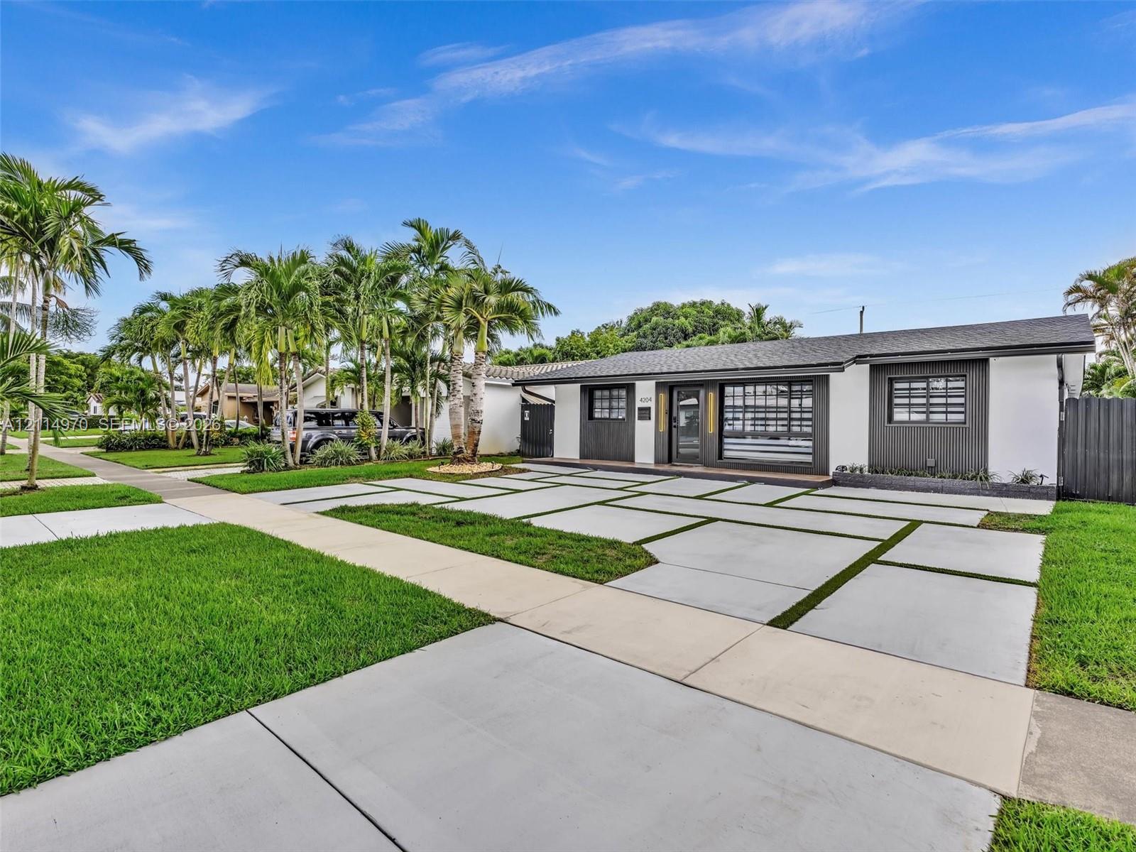 Single-story exterior featuring a grid-pattern concrete driveway, manicured lawn, mature palms, and dark-finished exterior paneling
