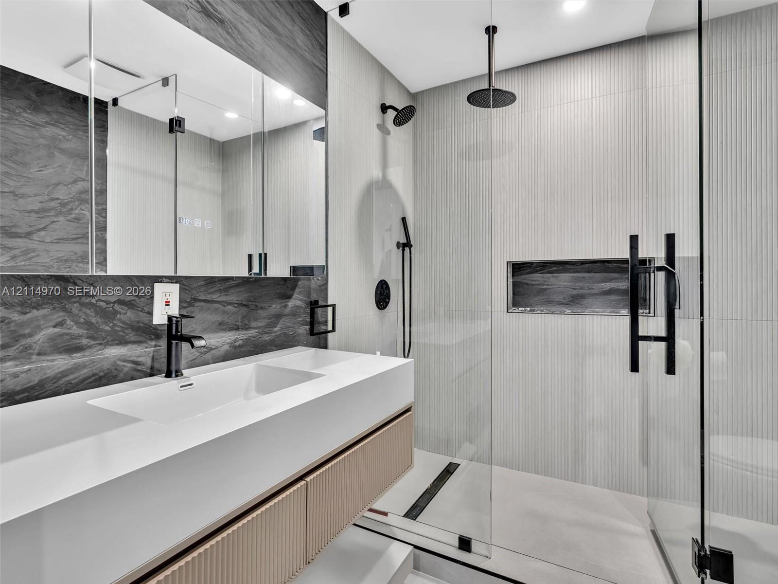 Floating vanity with a smooth solid-surface countertop and integrated sink, complemented by a recessed mirror, fluted-texture cabinetry, a glass-enclosed shower with a rainfall showerhead, and a built-in shower niche