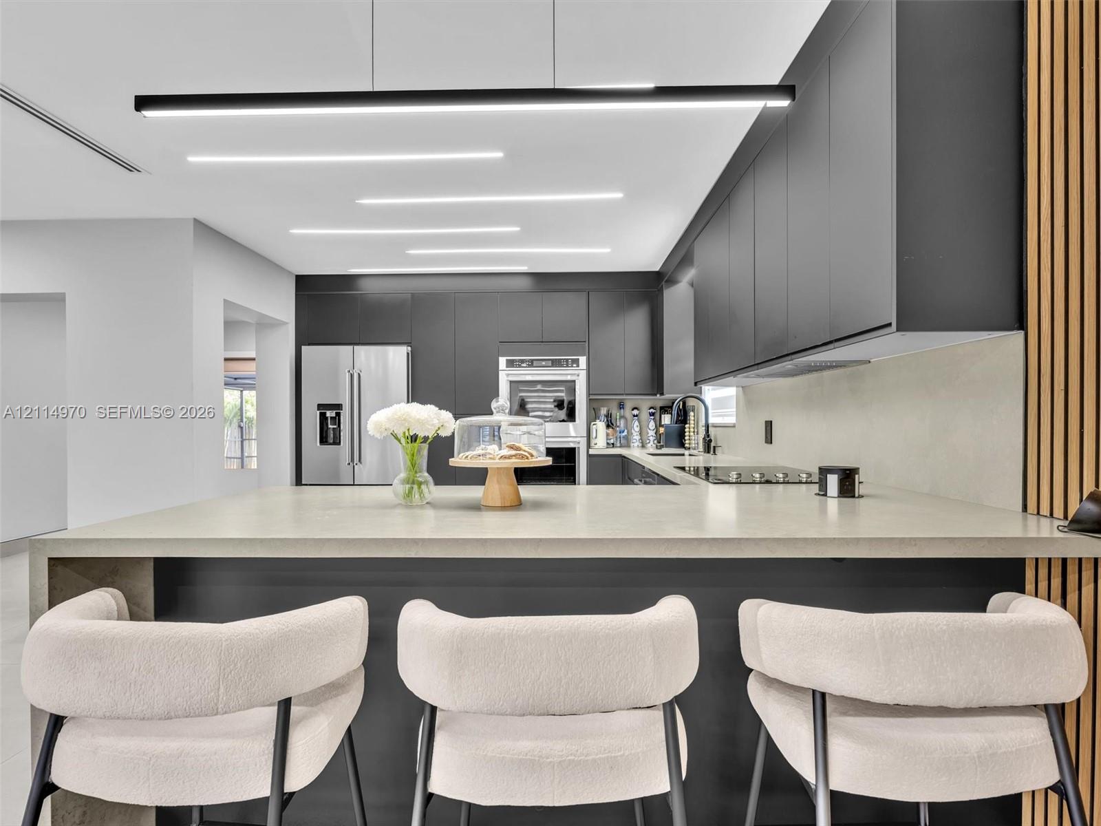 Modern kitchen featuring matte charcoal cabinetry, a large waterfall-edge island with a stone-finish countertop, built-in stainless steel appliances, recessed ceiling lighting, and decorative vertical wood-slat wall paneling Modern kitchen featuring matte charcoal cabinetry, a large waterfall-edge island with a stone-finish countertop, built-in stainless steel appliances, recessed ceiling lighting, and decorative vertical wood-slat wall paneling