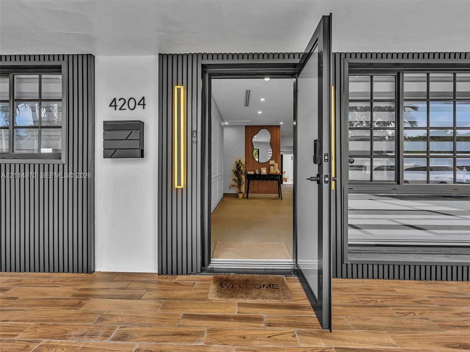Exterior entrance featuring vertical slat siding, wood-finish tile flooring, matte black grid-pane windows, and linear wall-mounted exterior lighting