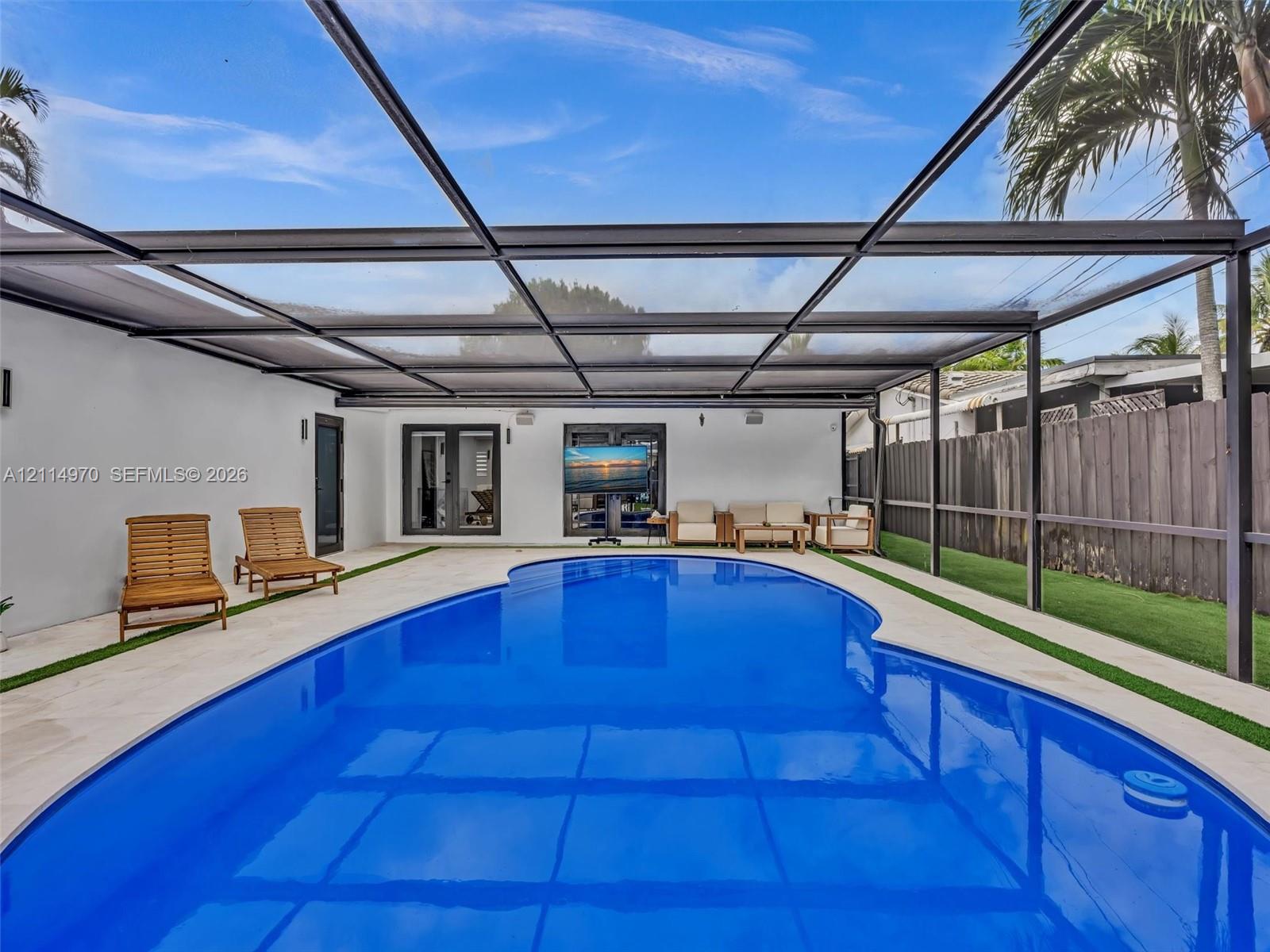 In-ground pool enclosed by a metal-framed screen structure, light-toned paved decking, artificial turf borders, and double-glass door entry