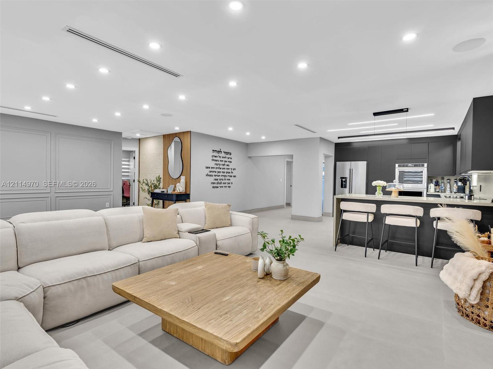 Open-concept interior featuring recessed lighting, matte black cabinetry, stone-surfaced kitchen island, wood-finish flooring, and wall paneling Open-concept interior featuring recessed lighting, matte black cabinetry, stone-surfaced kitchen island, wood-finish flooring, and wall paneling