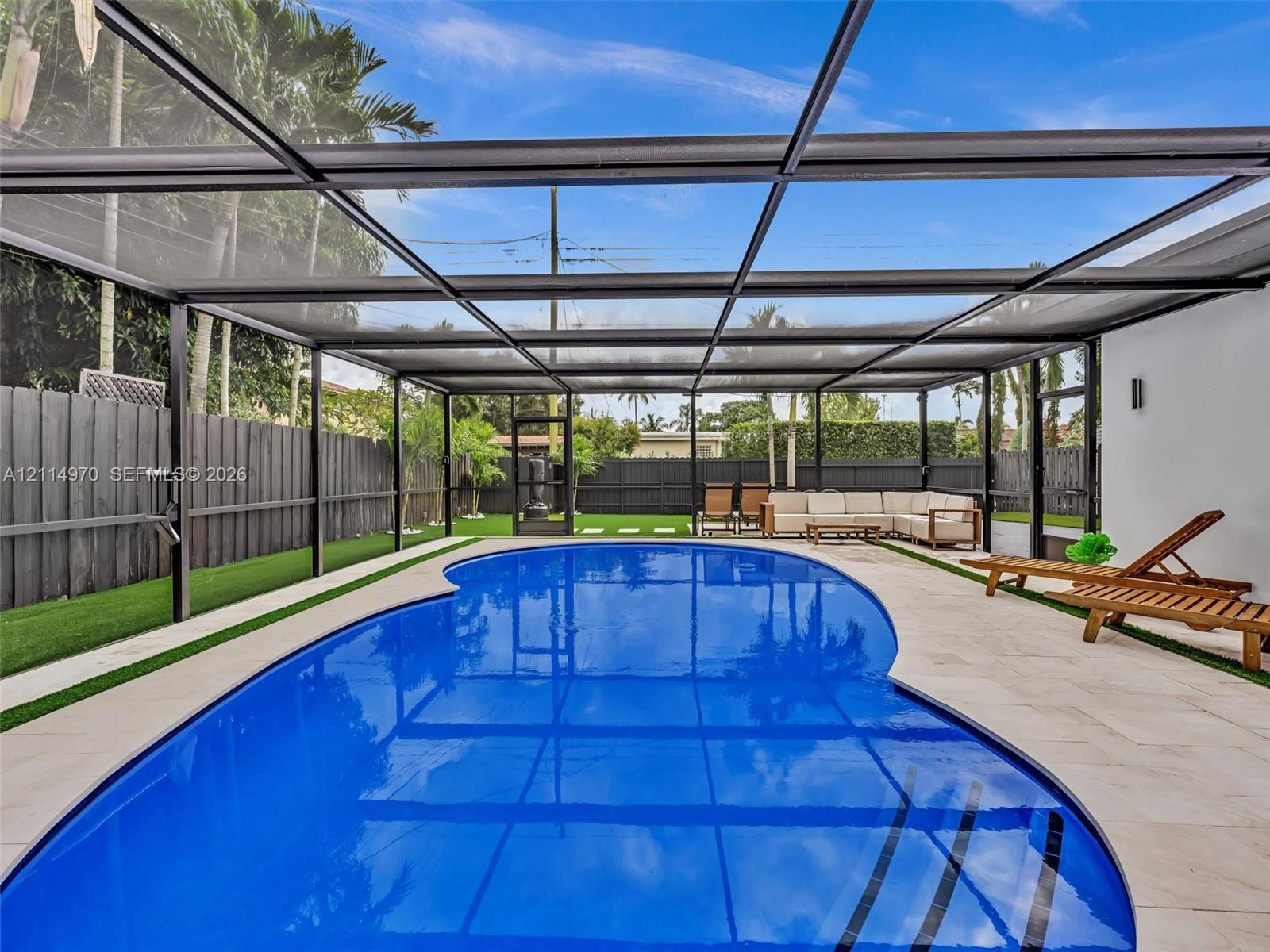 Screen-enclosed swimming pool featuring a curved concrete deck, synthetic turf perimeter, and dark wood-style privacy fencing