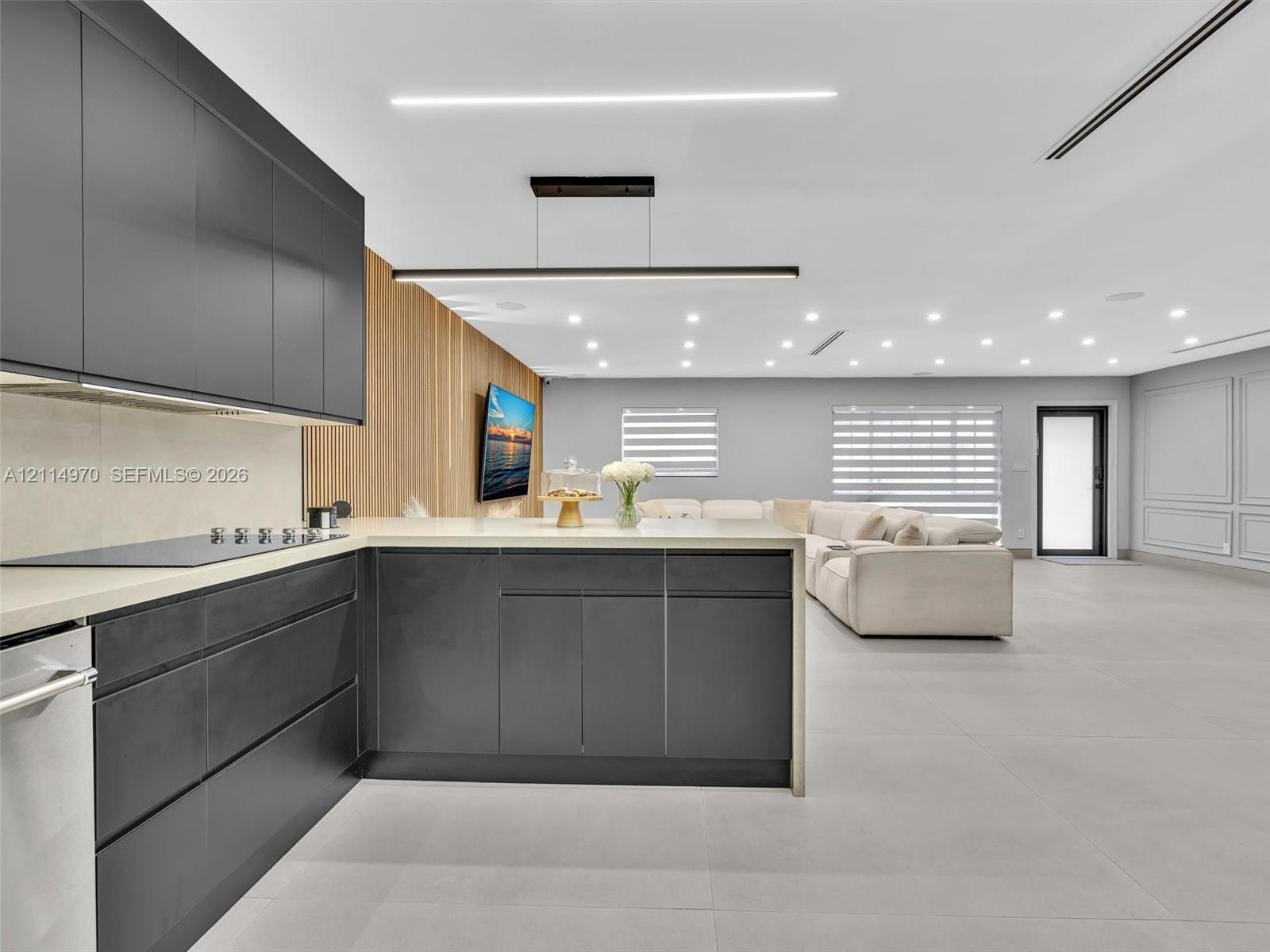 Modern kitchen featuring matte black slab-front cabinetry, light-toned stone countertops, integrated induction cooktop, slatted wood wall paneling, and large-format porcelain tile flooring Modern kitchen featuring matte black slab-front cabinetry, light-toned stone countertops, integrated induction cooktop, slatted wood wall paneling, and large-format porcelain tile flooring
