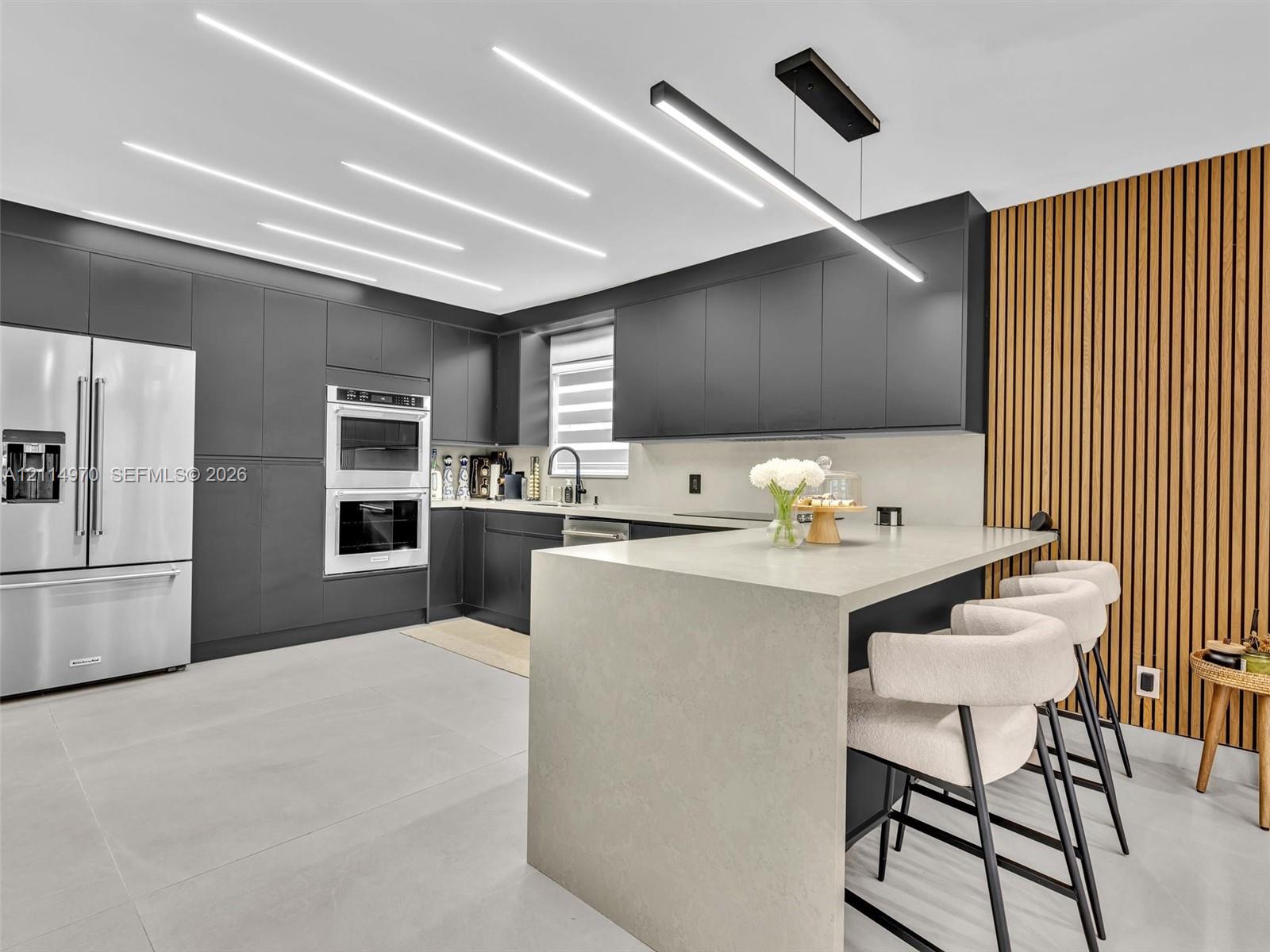Contemporary kitchen featuring matte charcoal cabinetry, a waterfall-edge stone island, linear recessed ceiling lighting, a wood-slat accent wall, and large-format tile flooring