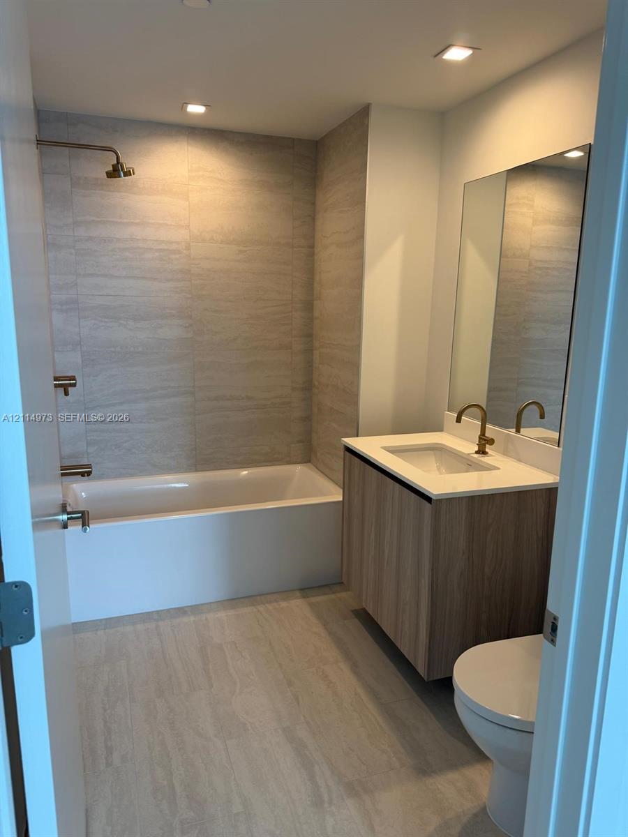 Bathroom featuring a floating vanity with wood-finish cabinetry, a white countertop with an integrated sink, a rectangular wall-mounted mirror, floor-to-ceiling large-format tile surround, and metallic fixtures