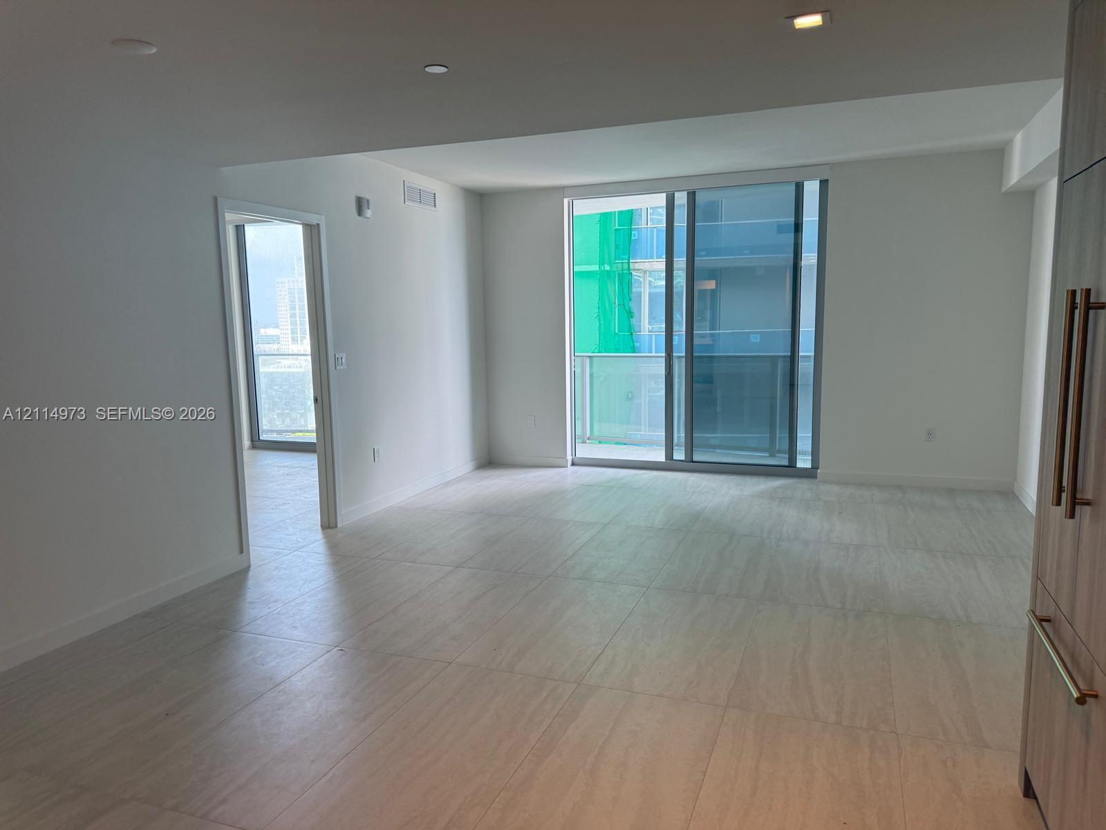 Open-concept living space featuring neutral large-format tile flooring, floor-to-ceiling sliding glass doors, built-in cabinetry with metallic pulls, and recessed lighting