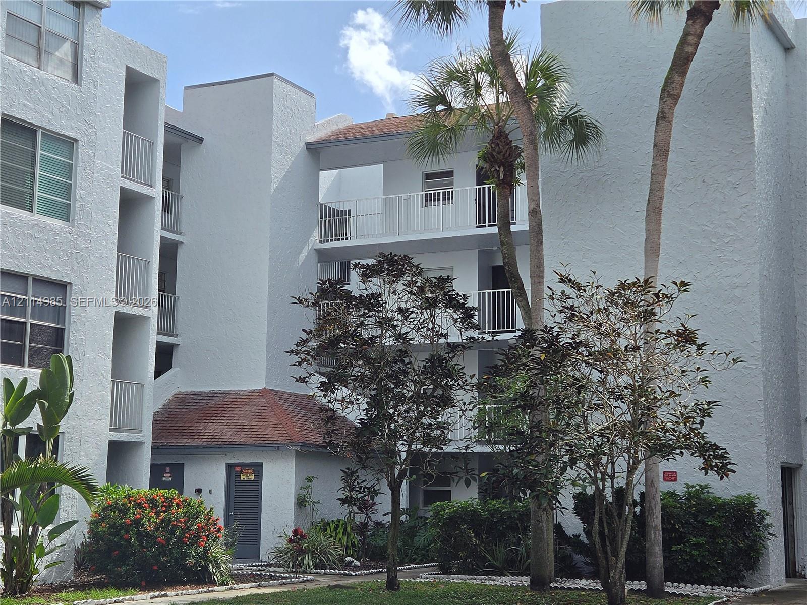Multi-story residential building exterior featuring a textured stucco facade, tiered balconies with white railings, and integrated landscaping with mature palm trees