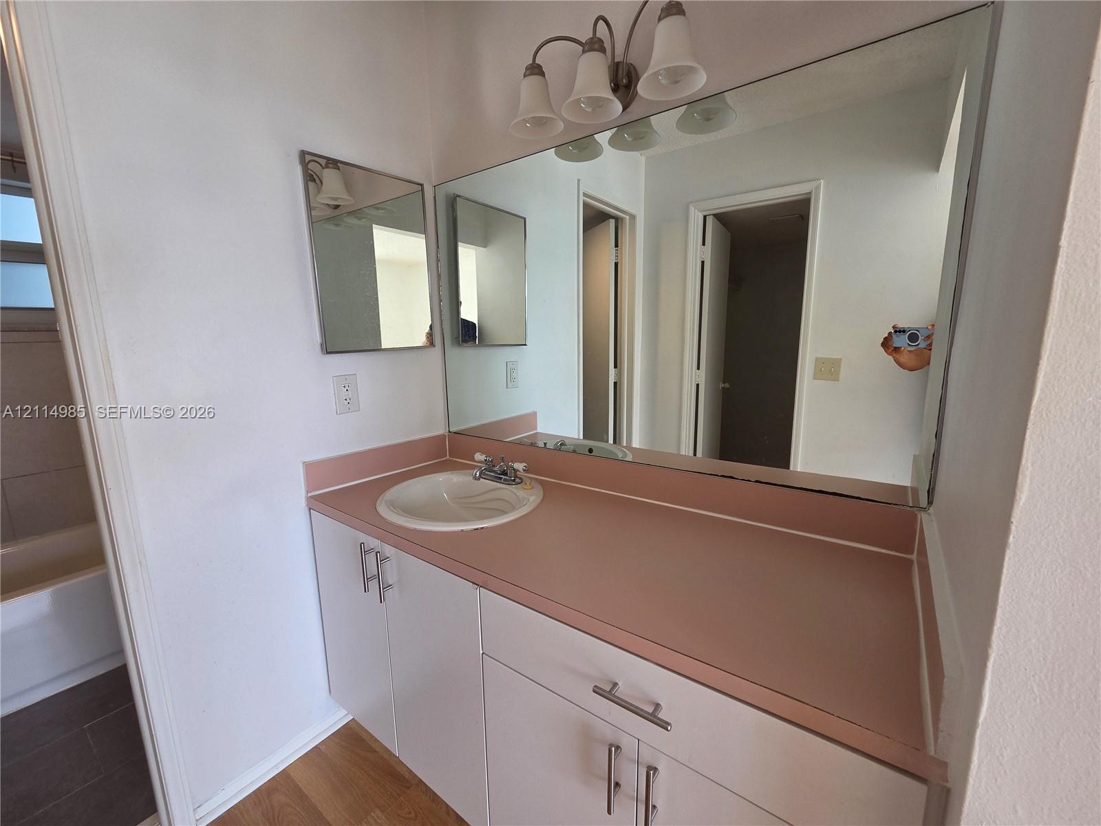Bathroom vanity featuring a solid-surface countertop, integrated basin, white cabinetry with brushed-metal hardware, and a large-format vanity mirror with a three-light fixture