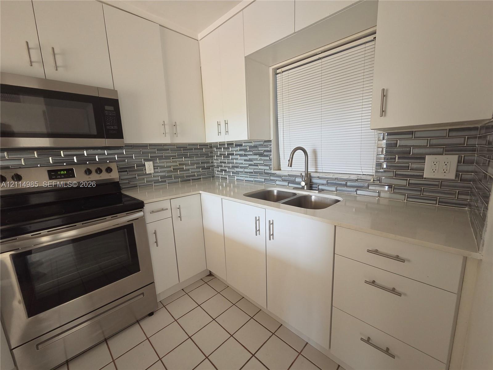 Kitchen featuring white flat-panel cabinetry, stainless steel appliances, light-toned stone countertops, a multi-tonal glass tile backsplash, and dual-basin sink with a gooseneck faucet