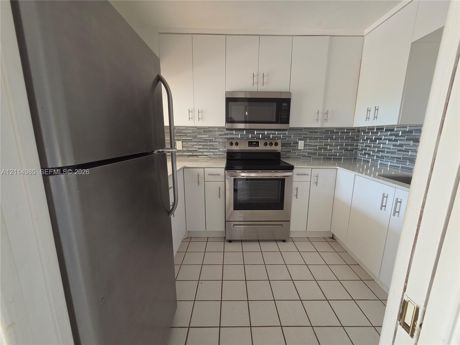 Kitchen featuring white shaker-style cabinetry, stainless steel appliances, patterned glass tile backsplash, stone-surface countertops, and tiled flooring