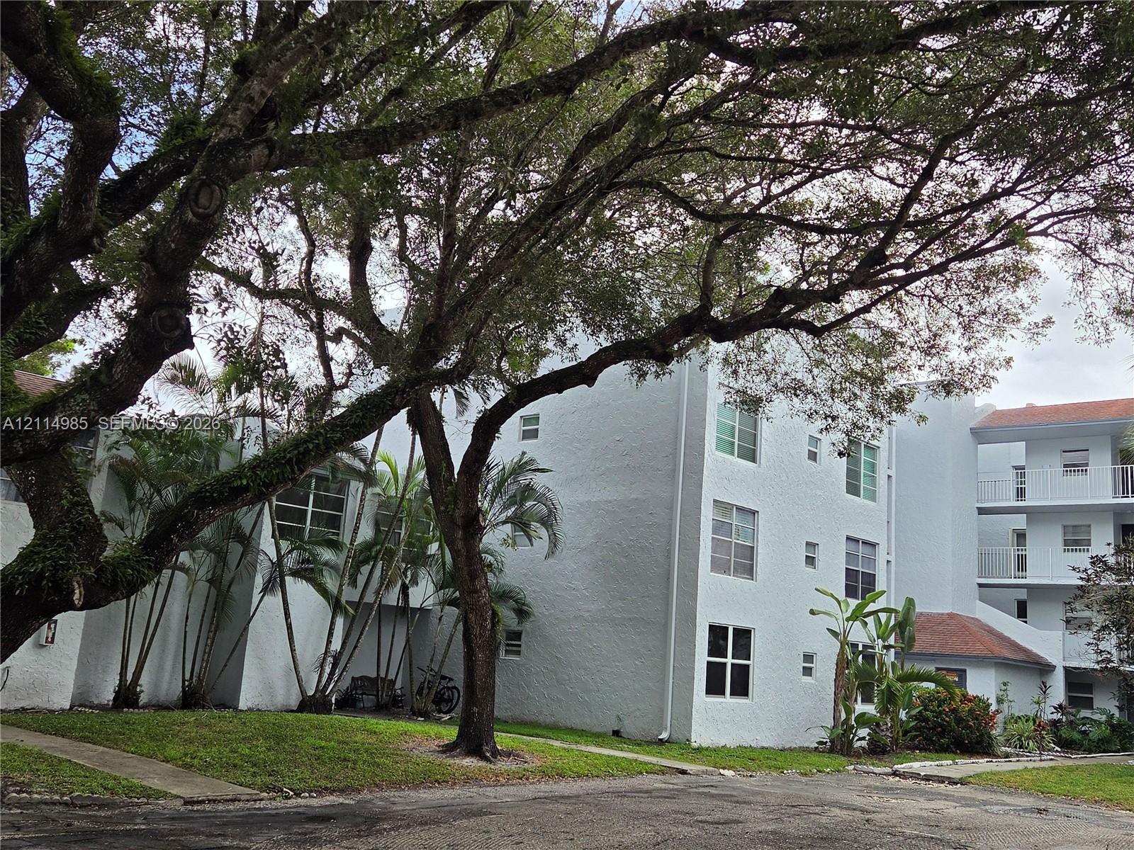 Multi-story building featuring textured stucco exterior, grid-pattern windows, multi-level exterior balconies with white railings, and mature trees