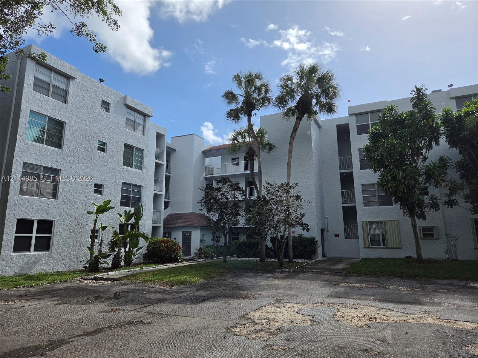 Multi-story residential building exterior featuring a textured stucco facade, asphalt parking area, and mature palm trees
