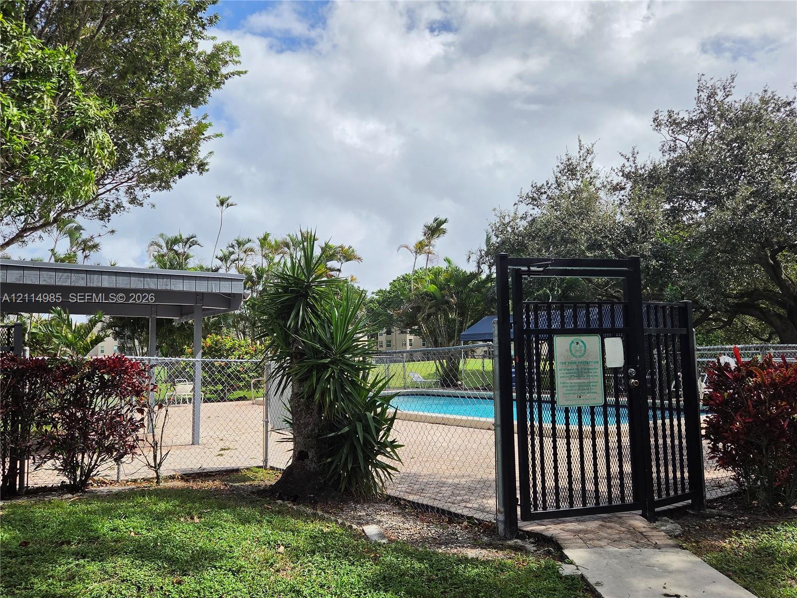 Outdoor swimming pool with paved decking, covered patio area, and metal security fencing surrounded by mature landscaping