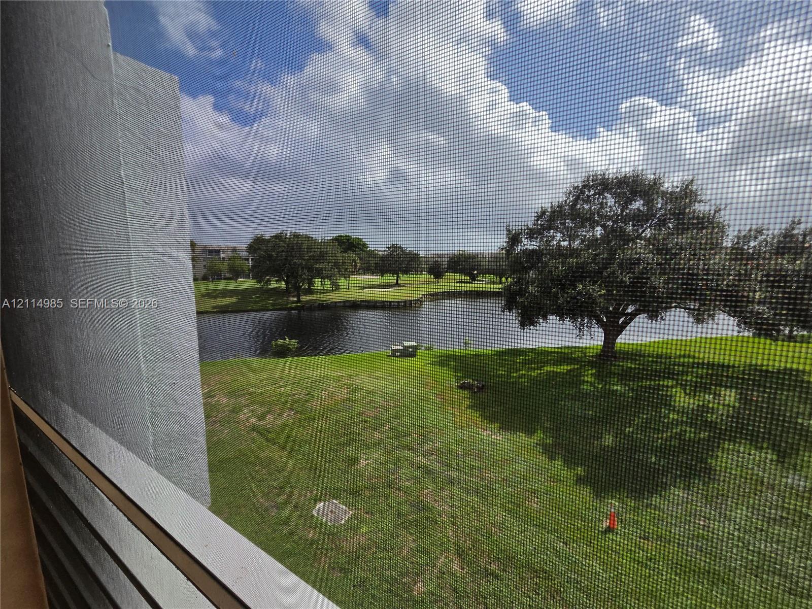 Exterior balcony featuring a white metal railing and a mesh screen overlooking a grass lawn, a pond, and mature trees