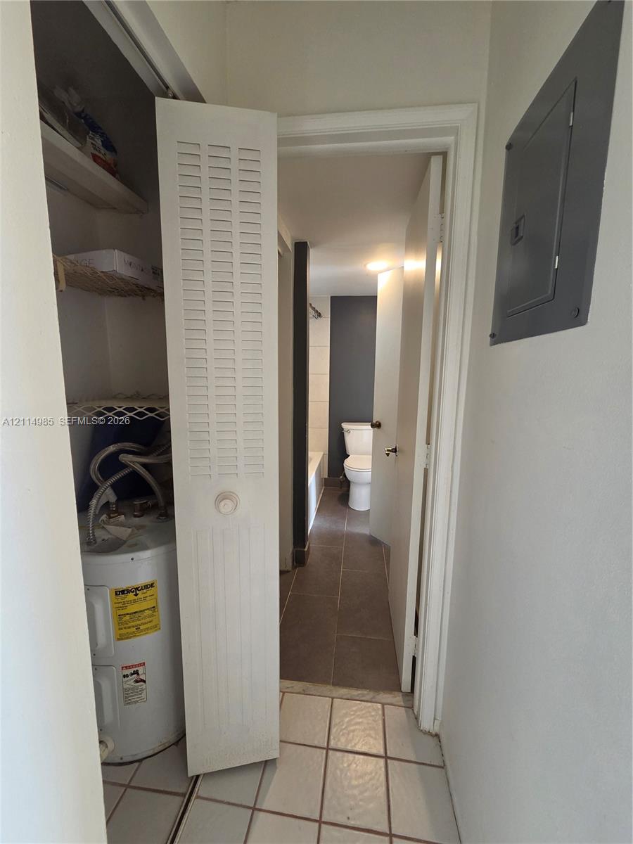 Utility closet featuring a louvered door, integrated shelving, and a water heater, with large-format tile flooring and an electrical service panel