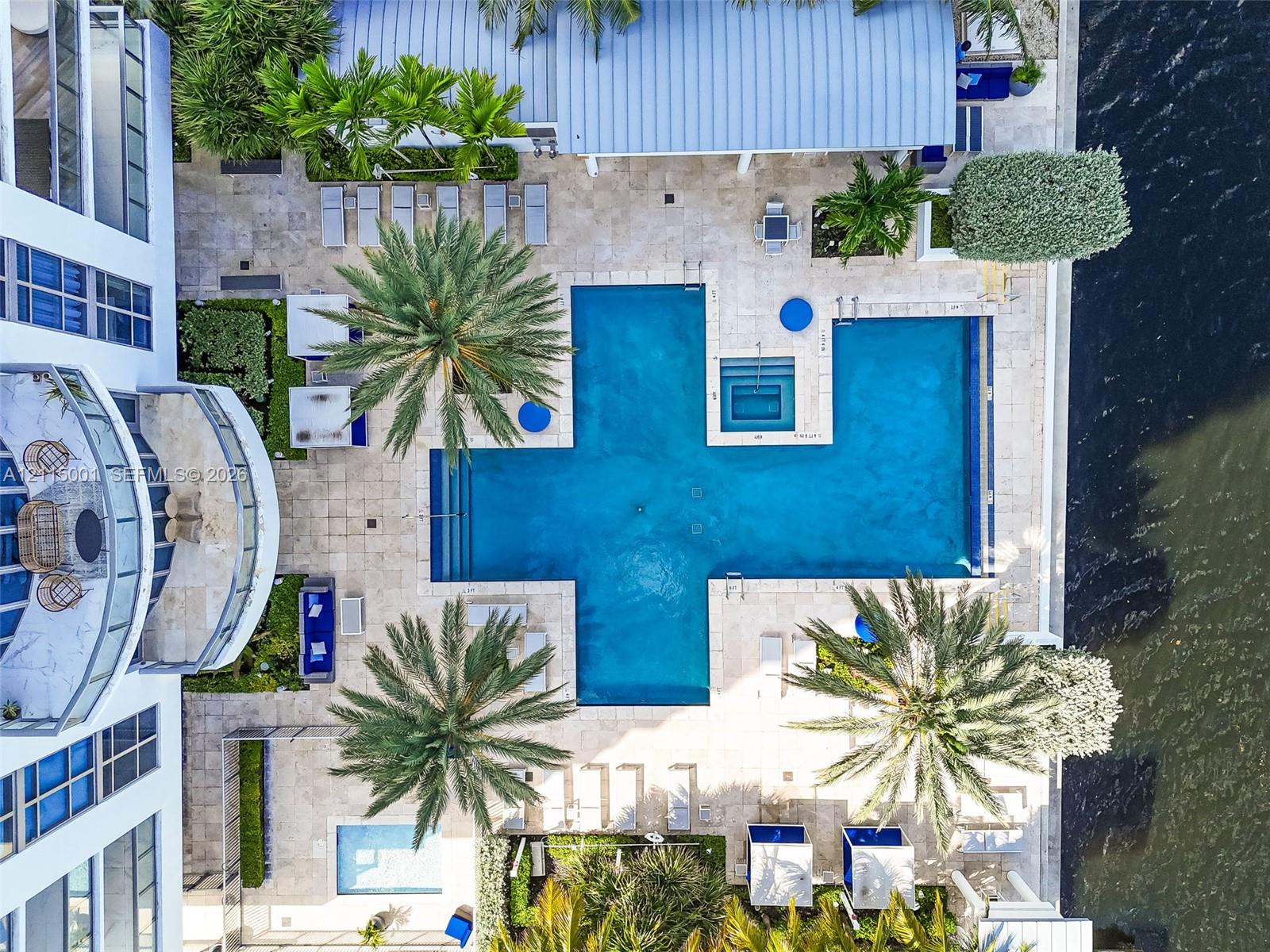 Waterfront swimming pool area featuring natural stone decking, integrated spa, mature palm landscaping, and metal-roofed covered terrace Waterfront swimming pool area featuring natural stone decking, integrated spa, mature palm landscaping, and metal-roofed covered terrace