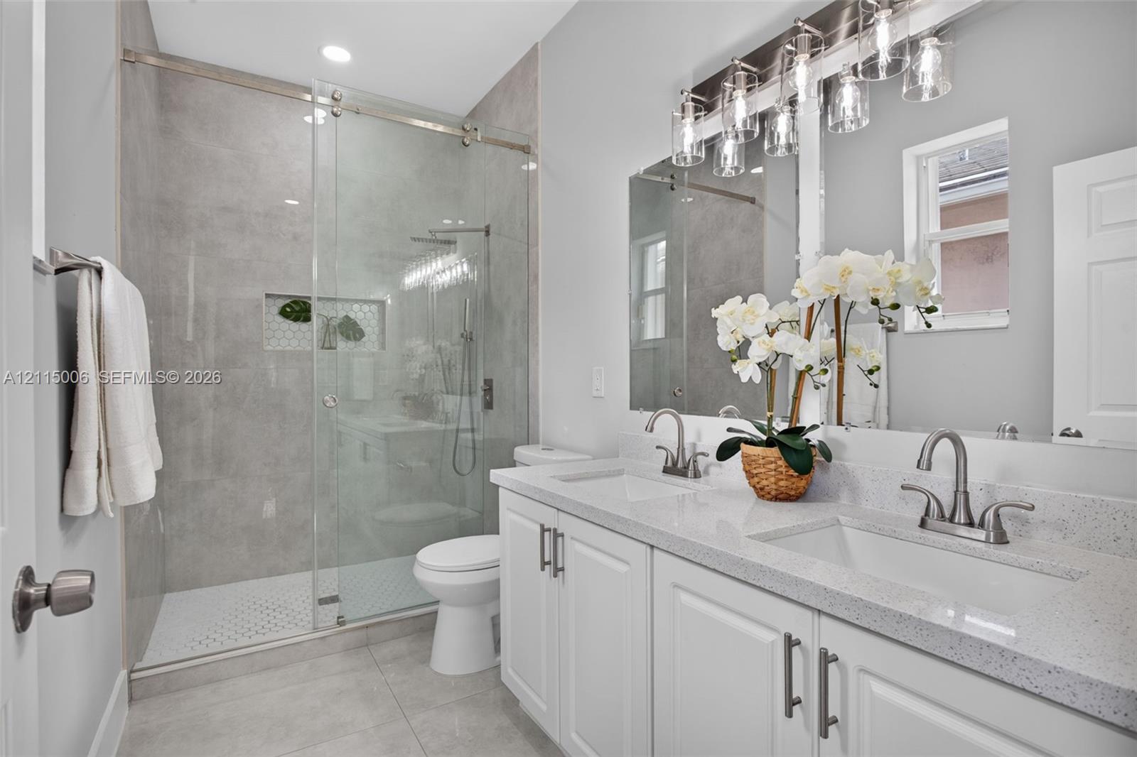 Double vanity with light-colored composite countertops, walk-in shower with glass enclosure and tiled surround, recessed shower niche, and multi-light vanity fixture