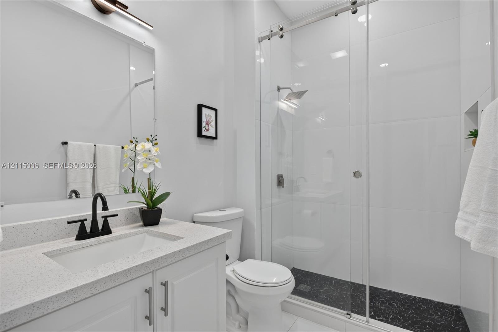 White cabinetry vanity with solid-surface countertop, matte black faucet, glass-enclosed shower with contrasting dark stone-finish flooring, and wall-mounted light fixture