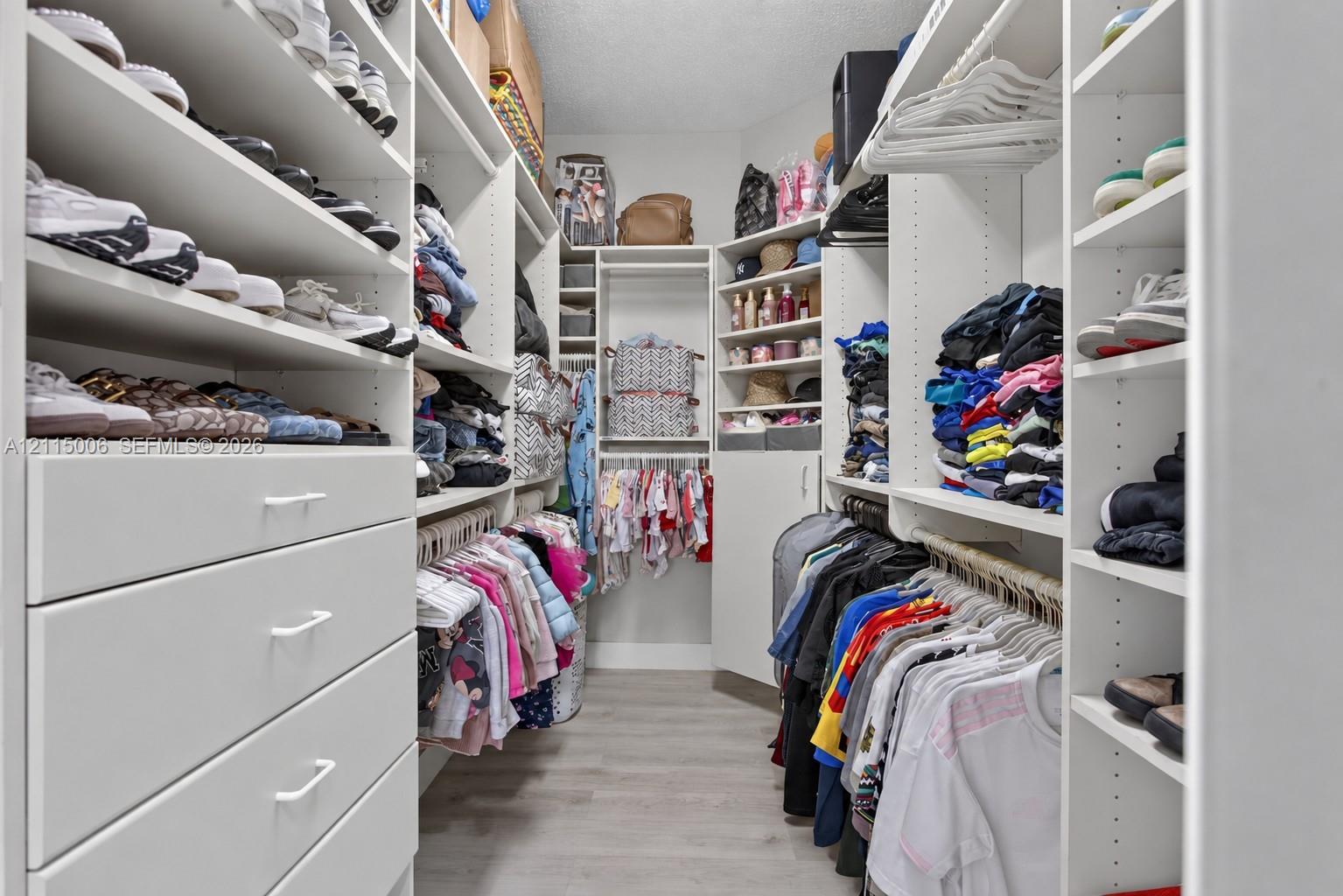Custom walk-in closet featuring white built-in shelving, multiple hanging rods, integrated storage drawers with bar pulls, and wood-finish flooring