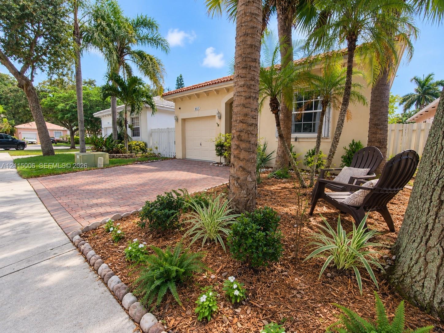 Stucco exterior featuring a tile roof, brick-paver driveway, single-car garage, and mulched landscaping with mature palm trees Stucco exterior featuring a tile roof, brick-paver driveway, single-car garage, and mulched landscaping with mature palm trees