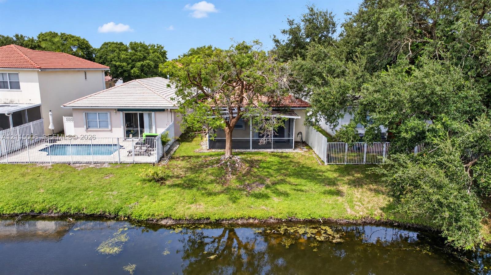 Waterfront exterior featuring a screen-enclosed patio, in-ground pool with concrete surround, perimeter white metal fencing, and mature landscaping
